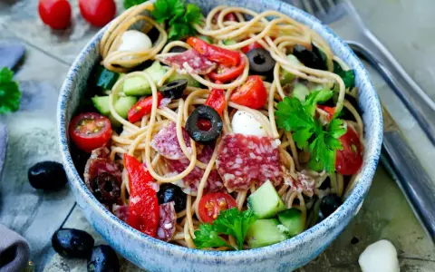 Bunter Spaghetti-Salat mit Salami, Tomaten, Oliven und Mozzarella in einer blauen Schüssel, garniert mit Petersilie.