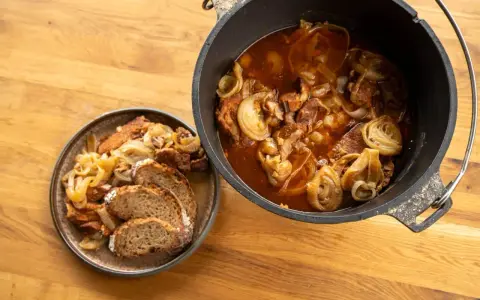 Herzhaftes Zwiebelfleisch im Dutch Oven, daneben eine Portion mit Brot auf einem Teller.