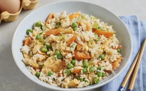 Schüssel mit gebratenem Eierreis wie beim Chinesen, garniert mit Erbsen und Karotten. Daneben liegen Stäbchen.