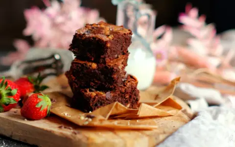 Saftige Brownies nach original Rezept, gestapelt auf einem Holzbrett mit Erdbeeren und einem Krug Milch.