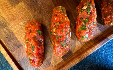 Rohe Cevapcici, bereit für das Rezept, liegen auf einem Holzbrett. Die Hackfleischröllchen sind mit Kräutern und Gewürzen verfeinert.