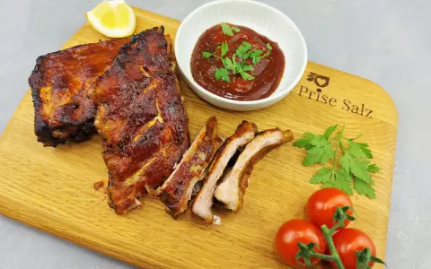 Butterzarte Spareribs im Backofen, glasiert und saftig, serviert mit BBQ-Sauce, Zitrone und frischen Tomaten auf einem Holzbrett.