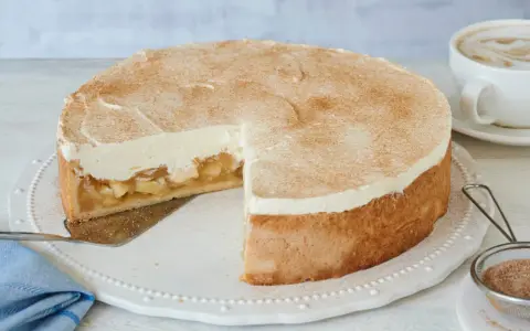 Saftige Apfel-Schmand-Torte mit Zimt bestäubt, ein Stück herausgeschnitten, daneben eine Tasse Cappuccino.