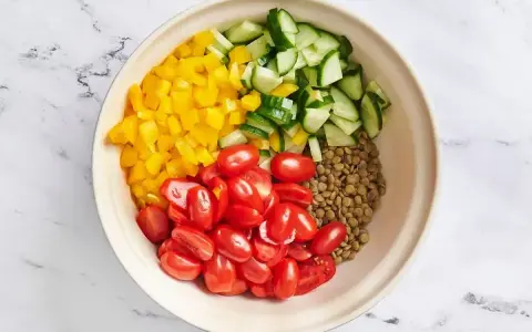 Zutaten für ein Linsensalat Rezept: gekochte Linsen, gewürfelte gelbe Paprika, Kirschtomaten und Gurkenstücke in einer Schüssel.