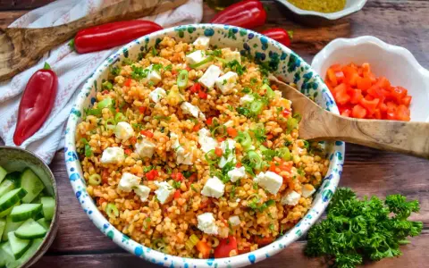Ein bunter Bulgursalat mit Feta, Paprika und Frühlingszwiebeln, garniert mit Petersilie. Dazu Gurkenscheiben und rote Peperoni.