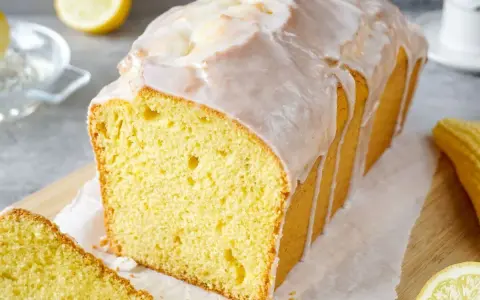 Saftiger Zitronenkuchen mit Zuckerguss, der aussieht wie der beste zitronenkuchen der welt. Ein Stück ist bereits angeschnitten.