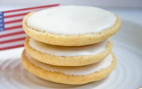 Drei amerikaner Kekse mit weißer Glasur auf einem Teller, im Hintergrund eine kleine US-Flagge. Ein leckeres amerikaner Rezept.