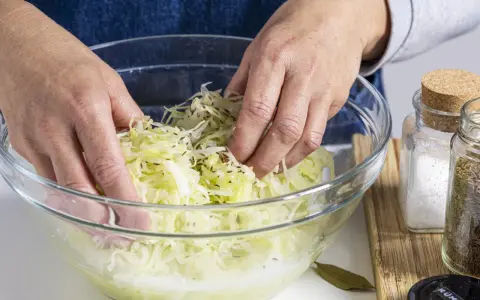 Hände mischen fein geschnittenen Weißkohl mit Kümmel in einer Glasschüssel. Ein Schritt im Sauerkraut Rezept.