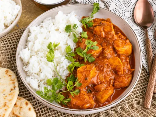 Ein Teller mit dampfendem Reis und cremigem Chicken Tikka Masala, garniert mit frischem Koriander. Dazu Naan-Brot und ein Löffel.