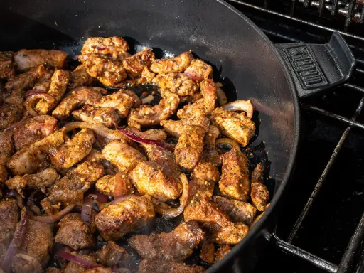 Würziges Gyros brutzelt in einem Dutch Oven auf dem Grill. Zarte Fleischstücke mit Zwiebeln garen in der Pfanne.