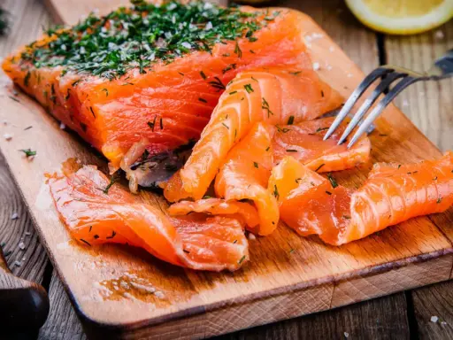 Frisch gebeizter Lachs mit Dill und Salz auf einem Holzbrett. Eine Gabel nimmt ein Stück Lachs auf.