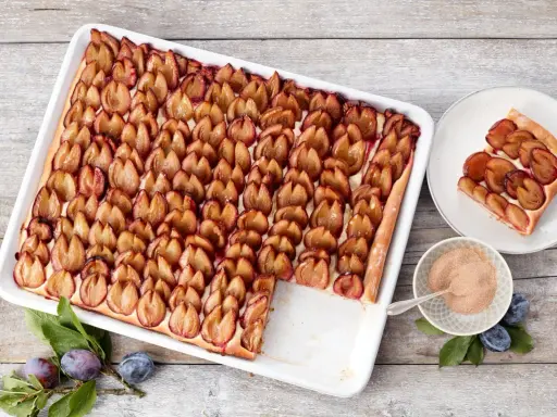 Ein köstlicher Zwetschgenkuchen vom Blech, mit saftigen Pflaumen belegt und einer Prise Zimt. Ein Stück ist schon auf einem Teller angerichtet.