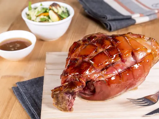 Glänzende Schweinshaxe sous vide auf Holzbrett, daneben Schüssel mit Sauce und Salat.