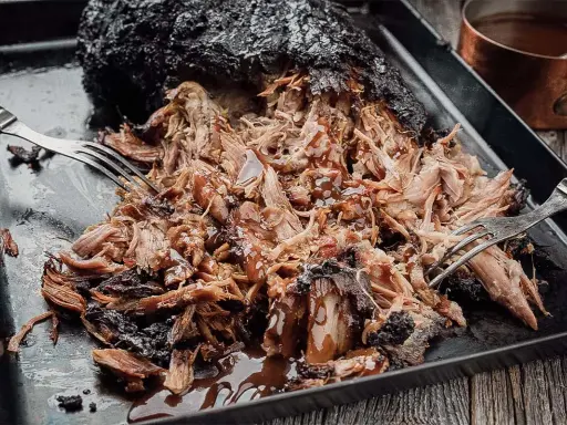 Saftiges Pulled Pork, frisch aus dem Smoker, wird mit Gabeln zerzupft und mit BBQ-Sauce beträufelt.