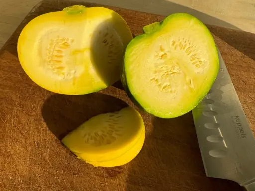 Zwei Hälften und ein Stück gelbe Zucchini liegen auf einem Holzbrett neben einem Messer.