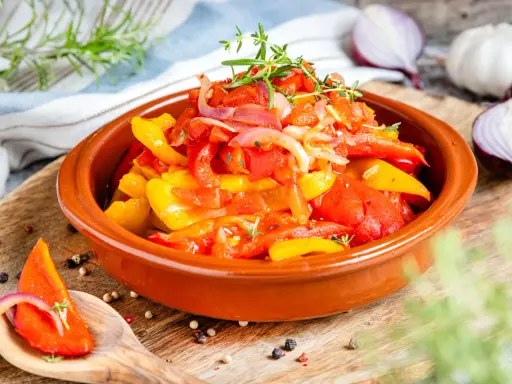 Buntes Peperonata-Gericht im Tontopf, garniert mit Kräutern. Ein Löffel zeigt ein Stück Paprika mit Zwiebeln, perfekt für das original Rezept.