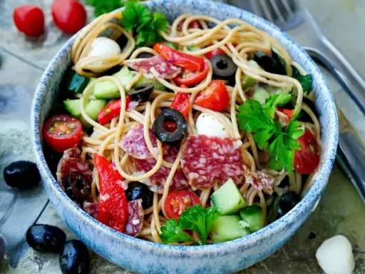 Bunter Spaghetti-Salat mit Salami, Tomaten, Oliven und Mozzarella in einer blauen Schüssel, garniert mit Petersilie.