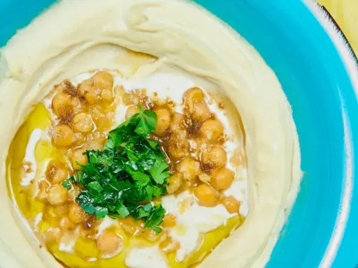 Cremiger Hummus, garniert mit Kichererbsen, Petersilie und Olivenöl. Ein authentisches Hummus Rezept original arabisch, serviert in einer blauen Schüssel.