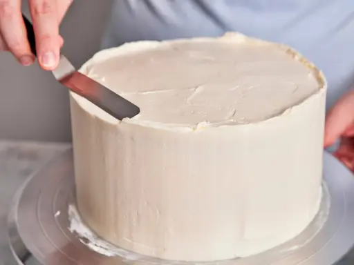 Eine Hand glättet mit einem Spachtel die cremige Buttercreme einer Torte, die für einen Geburtstag bestimmt ist.