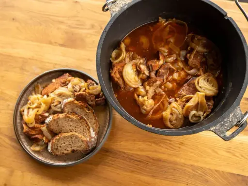 Herzhaftes Zwiebelfleisch im Dutch Oven, daneben eine Portion mit Brot auf einem Teller.