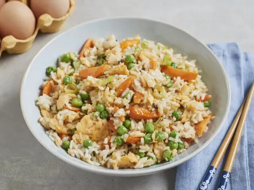 Schüssel mit gebratenem Eierreis wie beim Chinesen, garniert mit Erbsen und Karotten. Daneben liegen Stäbchen.