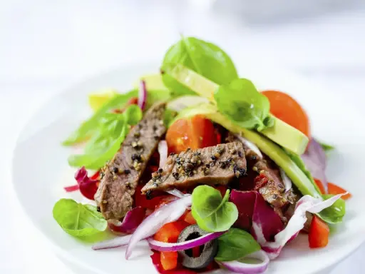 Frischer Salat mit zarten Rinderstreifen, Tomaten, roten Zwiebeln und Basilikum auf einem weißen Teller.