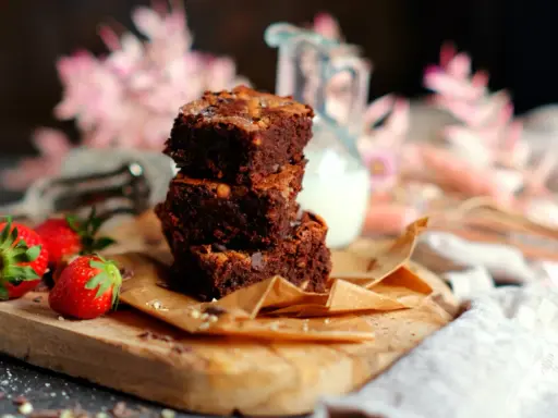 Saftige Brownies nach original Rezept, gestapelt auf einem Holzbrett mit Erdbeeren und einem Krug Milch.