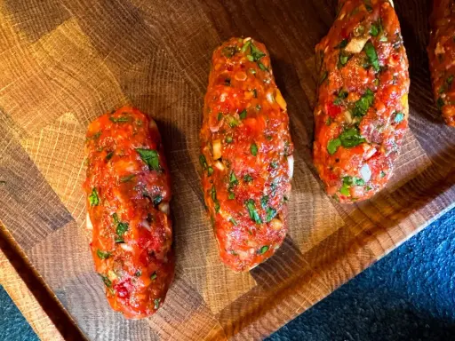 Rohe Cevapcici, bereit für das Rezept, liegen auf einem Holzbrett. Die Hackfleischröllchen sind mit Kräutern und Gewürzen verfeinert.