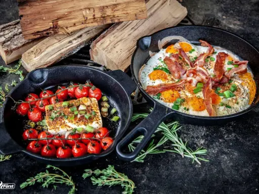 Zwei Gusseisenpfannen mit Eiern, Speck und Kirschtomaten mit Feta. Perfekt für low carb grillen.