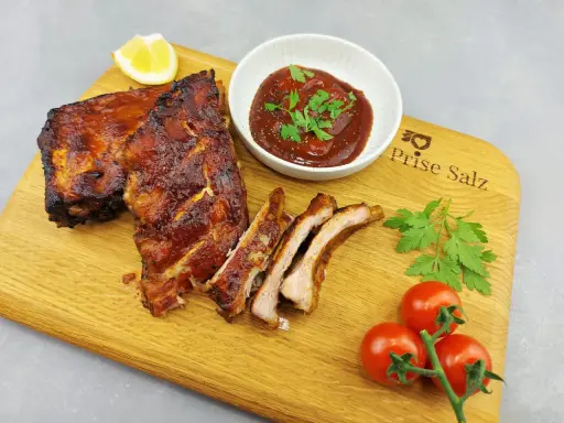 Butterzarte Spareribs im Backofen, glasiert und saftig, serviert mit BBQ-Sauce, Zitrone und frischen Tomaten auf einem Holzbrett.