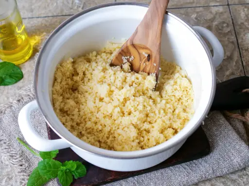 Ein Topf mit lockerem Couscous, bereit für ein marokkanisches Rezept. Ein Holzlöffel rührt die Körner auf. Daneben eine Flasche Öl und frische Minze.