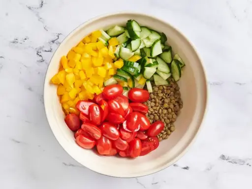 Zutaten für ein Linsensalat Rezept: gekochte Linsen, gewürfelte gelbe Paprika, Kirschtomaten und Gurkenstücke in einer Schüssel.