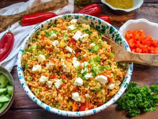Ein bunter Bulgursalat mit Feta, Paprika und Frühlingszwiebeln, garniert mit Petersilie. Dazu Gurkenscheiben und rote Peperoni.