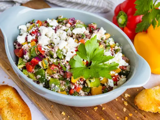 Bunter Paprikasalat mit Feta, garniert mit Petersilie, serviert in einer hellblauen Schüssel auf einem Holzbrett. Daneben liegen geröstete Brotscheiben.