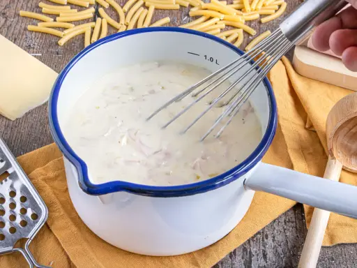 Cremige schinken sahne sauce wird in einem Topf mit einem Schneebesen zubereitet, umgeben von Nudeln, Käse und einer Reibe.