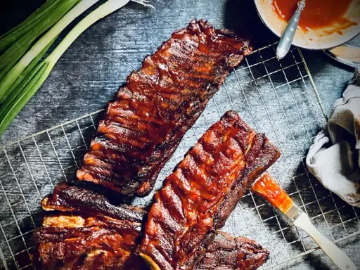 Saftige spareribs grillen holzkohle, mit BBQ-Sauce bestrichen, bereit zum Genuss. Grüne Frühlingszwiebeln und ein Pinsel liegen daneben.