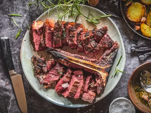 Saftiges, perfekt gegrilltes Bistecca alla Fiorentina, aufgeschnitten und mit Rosmarin garniert, daneben knusprige Kartoffeln.