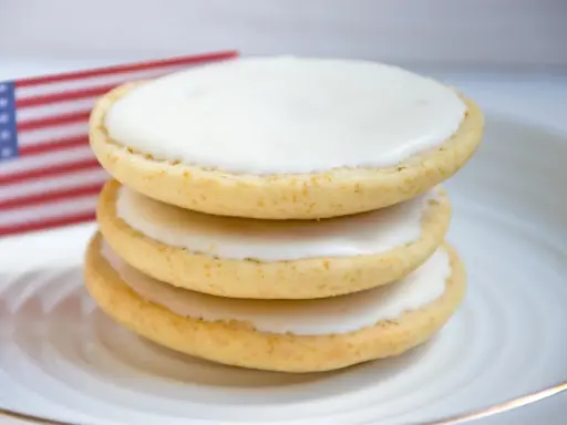 Drei amerikaner Kekse mit weißer Glasur auf einem Teller, im Hintergrund eine kleine US-Flagge. Ein leckeres amerikaner Rezept.