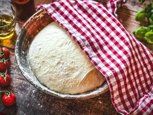 Pizzateig original rezept von einem italienischen gastronomen ruht in einer Schüssel, bedeckt mit einem rot-weißen Tuch. Tomaten und Olivenöl daneben.