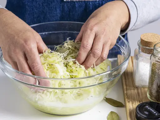 Hände mischen fein geschnittenen Weißkohl mit Kümmel in einer Glasschüssel. Ein Schritt im Sauerkraut Rezept.