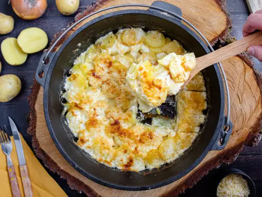 Cremige Kartoffeln im Dutch Oven, goldbraun gebacken. Eine Hand hebt mit einem Holzlöffel eine Portion heraus.