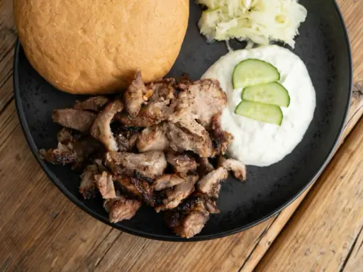 Saftiges Gyros vom Grill mit Tzatziki, Krautsalat und einem Brötchen auf schwarzer Platte.