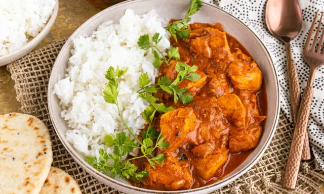 Ein Teller mit dampfendem Reis und cremigem Chicken Tikka Masala, garniert mit frischem Koriander. Dazu Naan-Brot und ein Löffel.