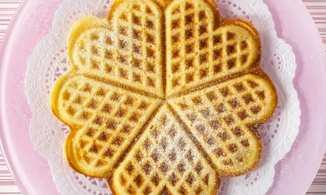 Herzförmige Waffeln ohne Ei, bestäubt mit Puderzucker, auf einem rosa Teller mit Spitzendeckchen.