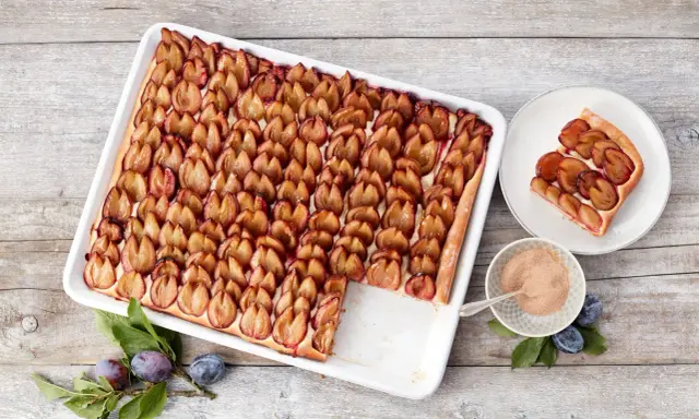 Ein köstlicher Zwetschgenkuchen vom Blech, mit saftigen Pflaumen belegt und einer Prise Zimt. Ein Stück ist schon auf einem Teller angerichtet.