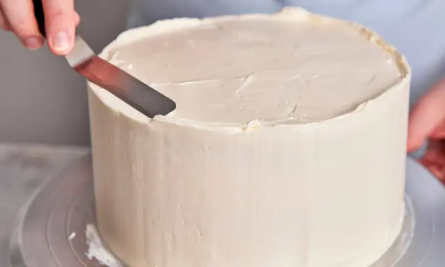 Eine Hand glättet mit einem Spachtel die cremige Buttercreme einer Torte, die für einen Geburtstag bestimmt ist.