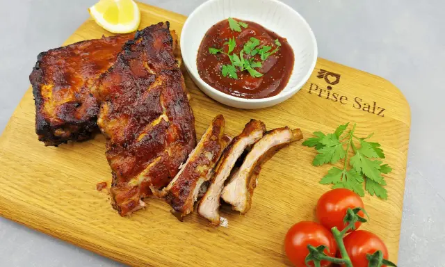 Butterzarte Spareribs im Backofen, glasiert und saftig, serviert mit BBQ-Sauce, Zitrone und frischen Tomaten auf einem Holzbrett.