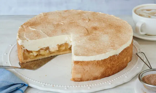Saftige Apfel-Schmand-Torte mit Zimt bestäubt, ein Stück herausgeschnitten, daneben eine Tasse Cappuccino.