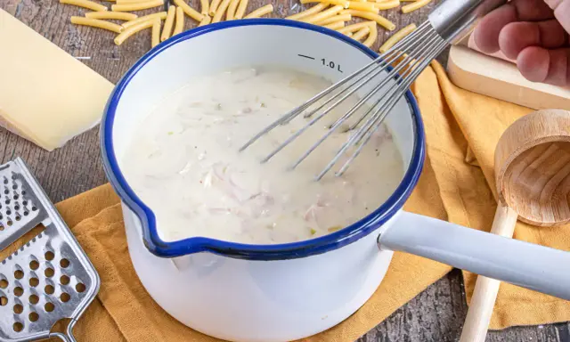 Cremige schinken sahne sauce wird in einem Topf mit einem Schneebesen zubereitet, umgeben von Nudeln, Käse und einer Reibe.