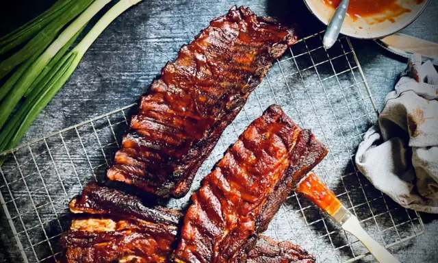 Saftige spareribs grillen holzkohle, mit BBQ-Sauce bestrichen, bereit zum Genuss. Grüne Frühlingszwiebeln und ein Pinsel liegen daneben.
